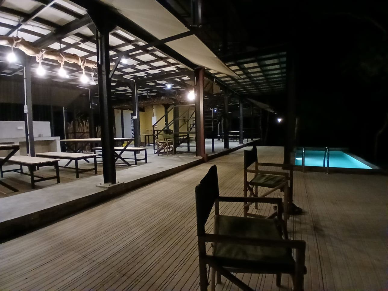 The long covered deck at night, chairs along it and the pool lit at the far end
